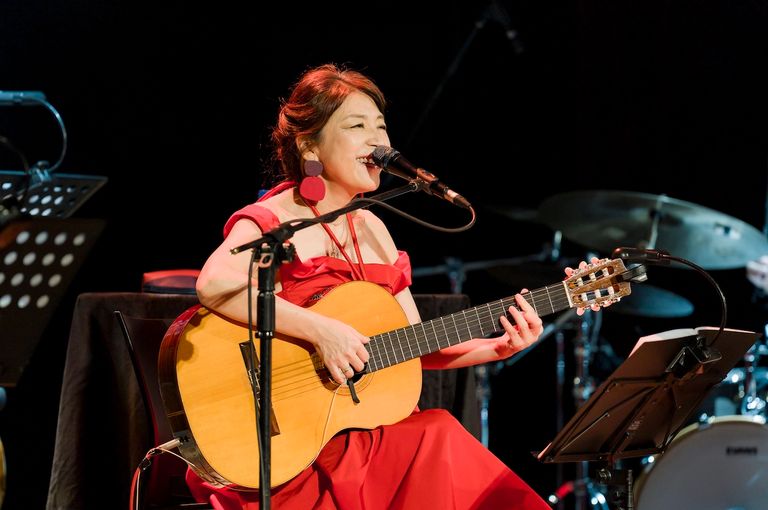 Bossa nova singer Lisa Ono performs live in a red dress, singing into a microphone while playing an acoustic guitar on stage.